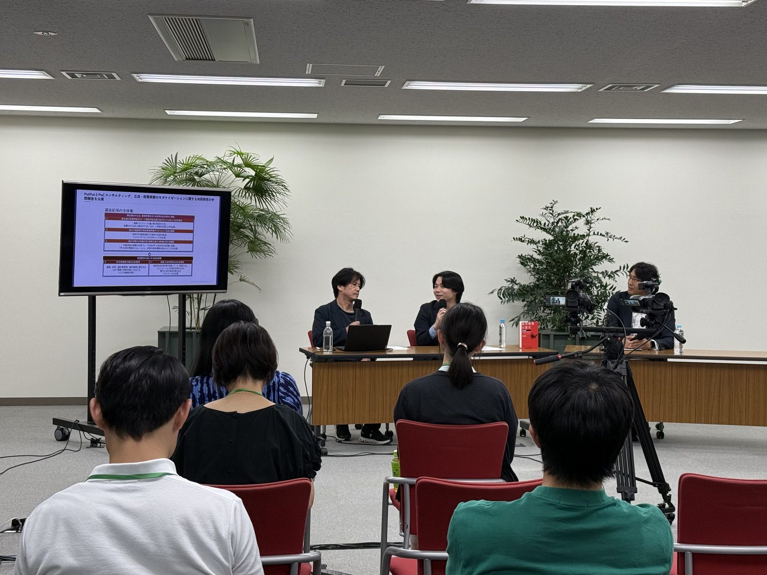 会場（官民共創HUB＠虎ノ門）の様子。前方に登壇者、後方に来場者、右手に配信用カメラ。ハイブリッド開催。