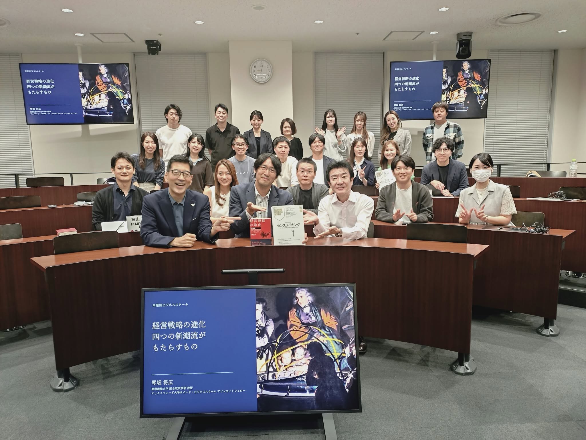 早稲田大学ビジネススクールの教室で、セッション後に撮影した集合写真。受講生と教員が段状の席に並び、前列では『経営戦略の進化〈理論編〉』と『DIAMOND ハーバード・ビジネス・レビュー』2026年10月号が掲げられている。手前と後方のモニターには、講演の表紙スライド「経営戦略の進化 四つの新潮流がもたらすもの」が映っている。
